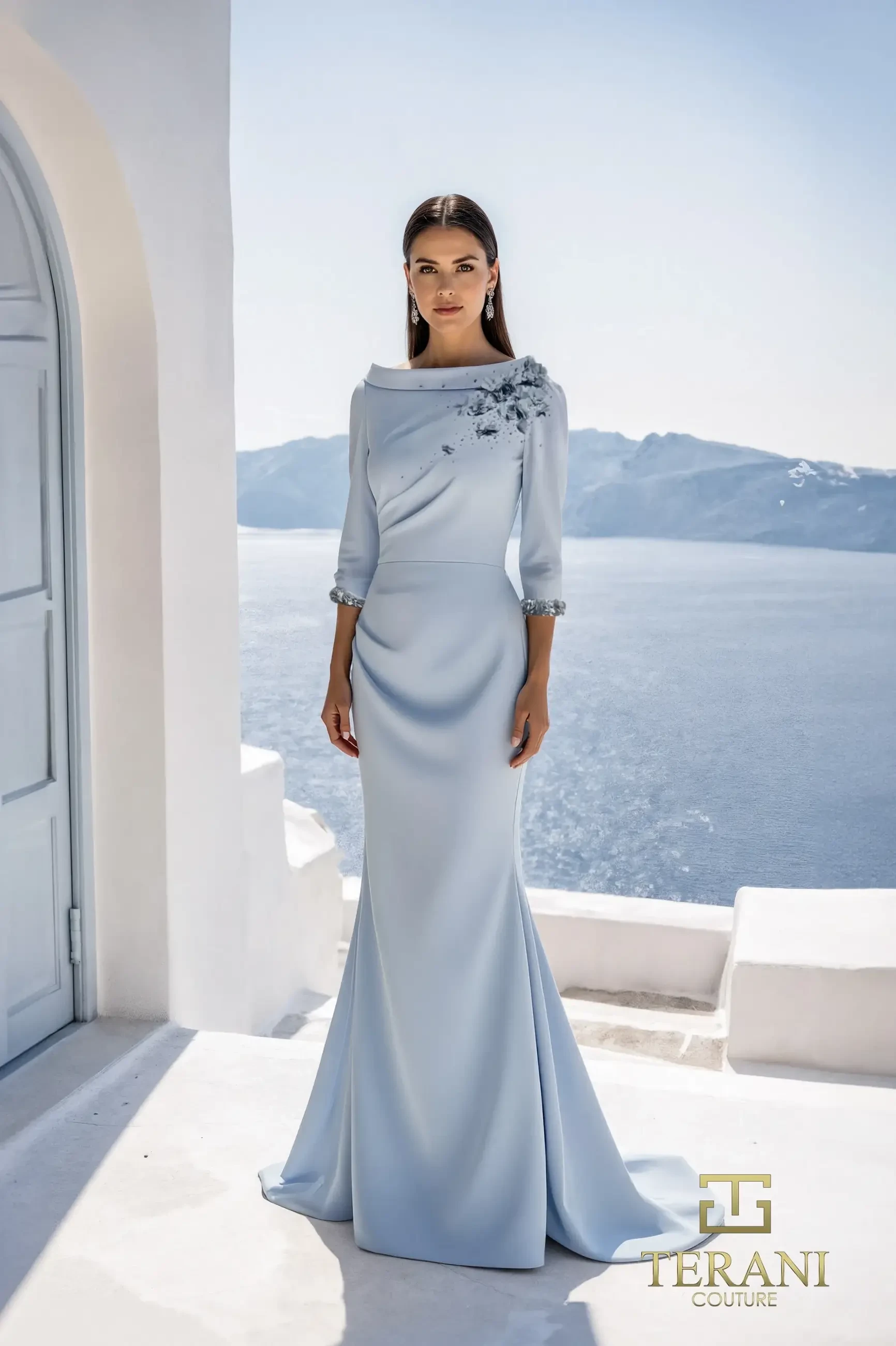 A woman in a light blue evening gown with three-quarter sleeves stands outdoors, showcasing a scenic backdrop of mountains and clear sky. The dress features floral embellishments on one shoulder.