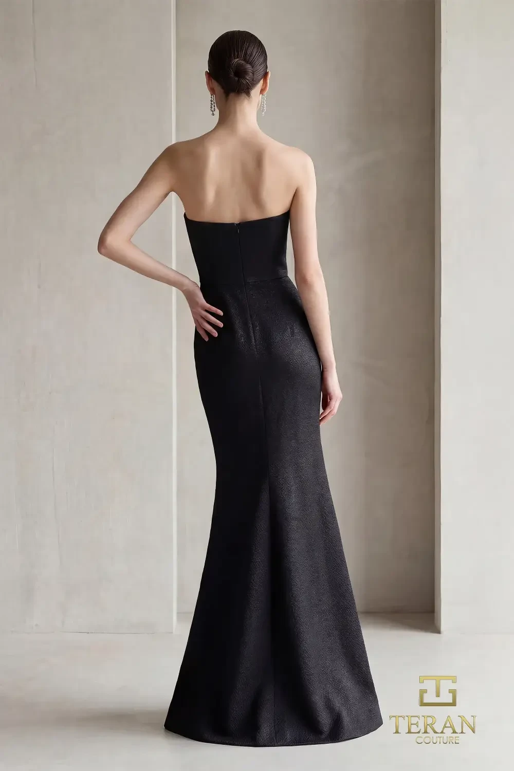 A model wearing a one-shoulder black gown with a fitted silhouette and decorative detailing at the shoulder.