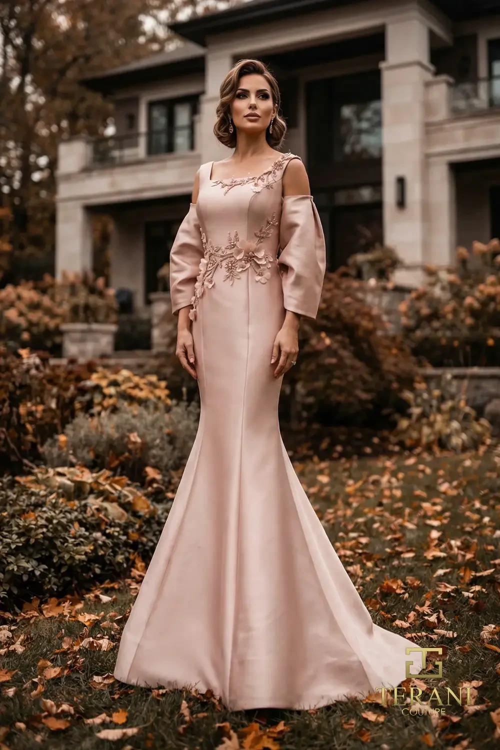 A woman in a formal, blush pink gown stands gracefully outside a modern house surrounded by autumn foliage.
