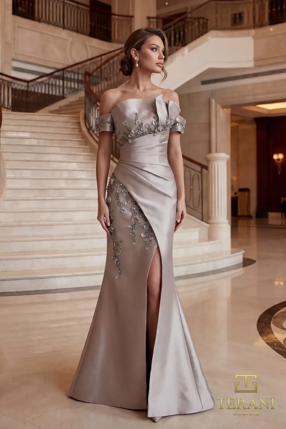 A woman in a sophisticated off-the-shoulder gown with embellishments, standing elegantly in a luxurious interior with a grand staircase in the background.