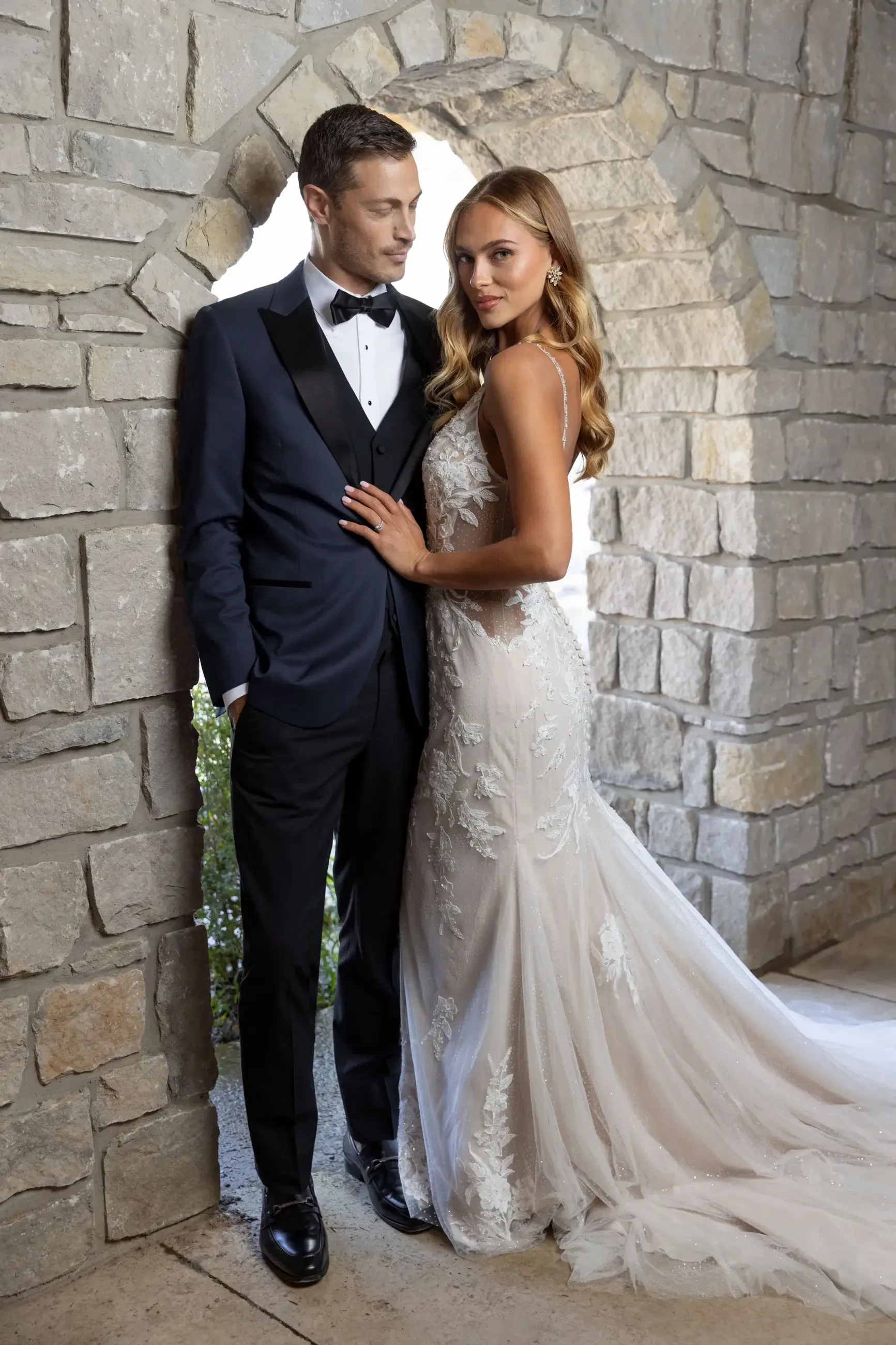 A couple poses together in formal attire, with the man wearing a tuxedo and the woman in a wedding gown. They are standing in front of a stone archway.