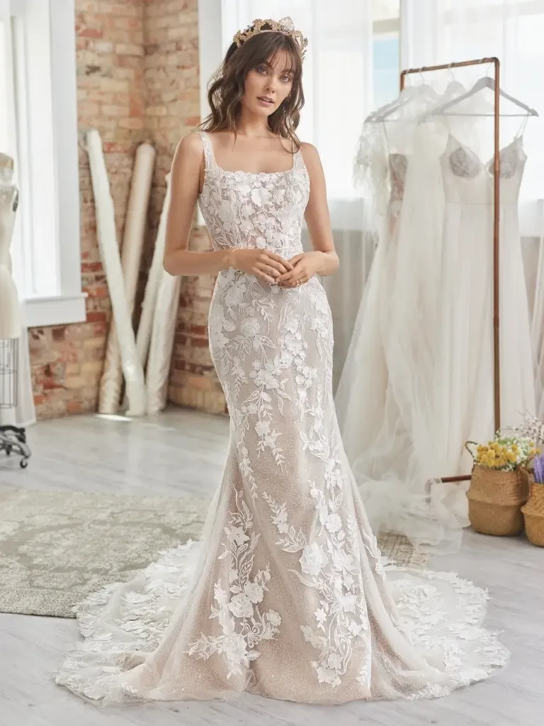A bride is standing in a bright room wearing an elegant, fitted wedding dress with floral lace details. The dress has thin straps and a train that flows behind her. She has her hair down with soft waves and is accessorized with a delicate flower crown. In the background, there are racks of bridal gowns and sewing materials.
