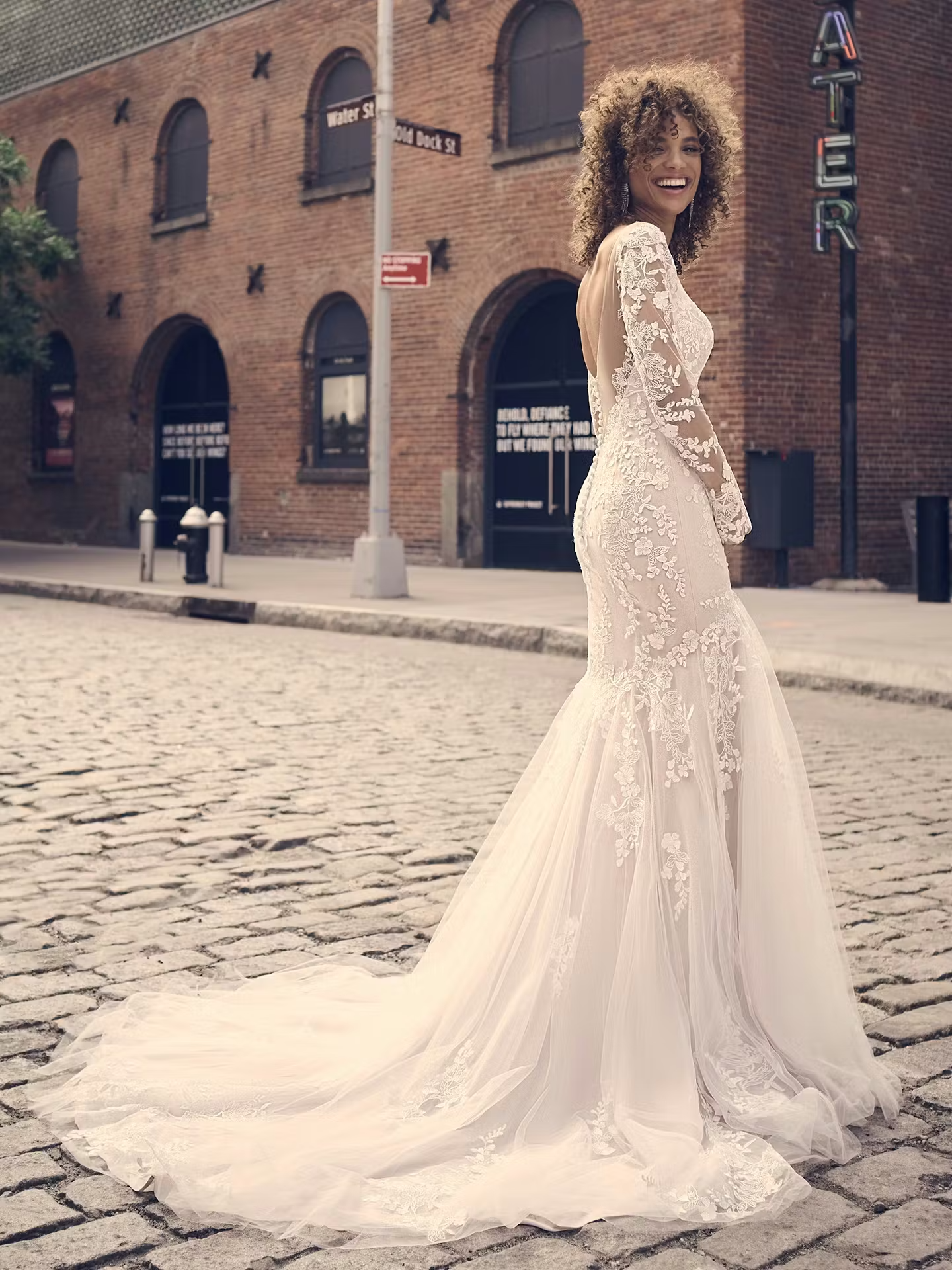 A woman wearing a long-sleeved wedding dress with lace details stands in a historic street, smiling confidently. The background features brick buildings and signs, creating a charming urban setting.