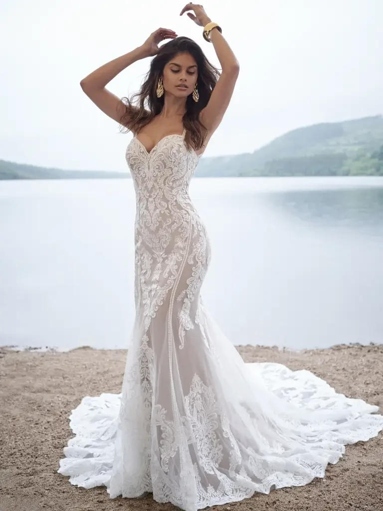A woman wearing an elegant, fitted lace wedding dress stands on a sandy shore near a body of water, with mountains in the background and her arms raised gracefully.