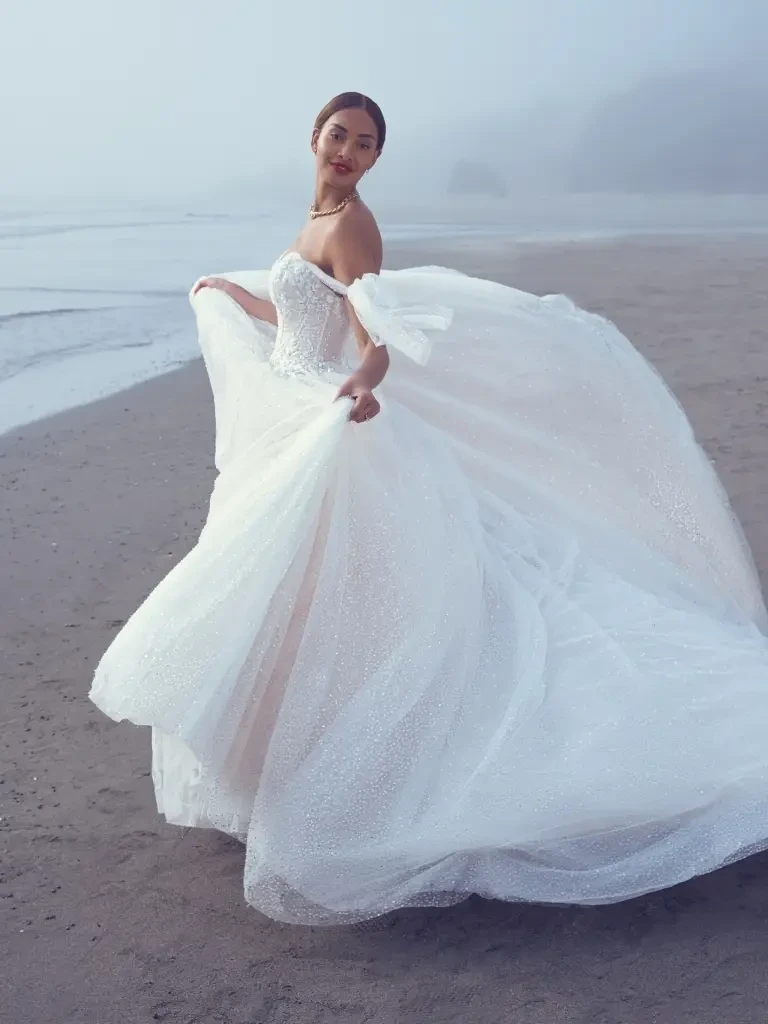 A woman in a flowing white wedding dress twirls on a beach with a foggy background.