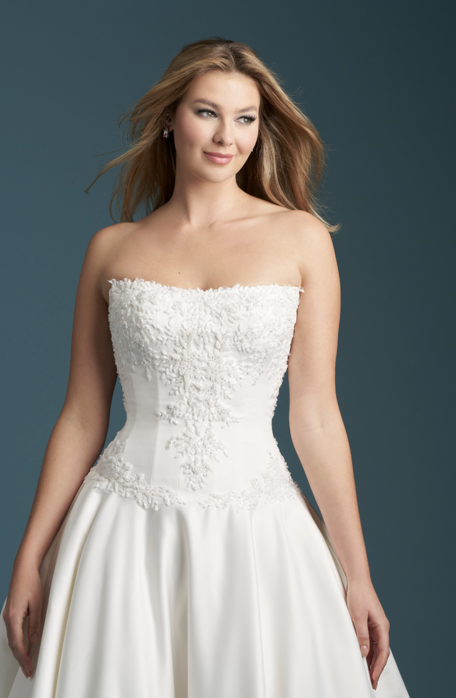 A woman in a strapless white wedding dress featuring intricate floral lace detail on the bodice. Her hair is loose, and the background is a soft teal.