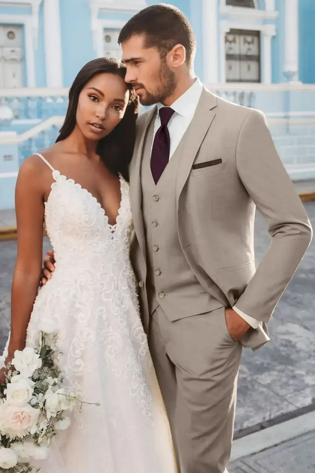 A couple posing together, the woman in a white wedding dress with lace details and the man in a gray suit with a vest and tie, standing outdoors. The woman holds a bouquet of flowers.