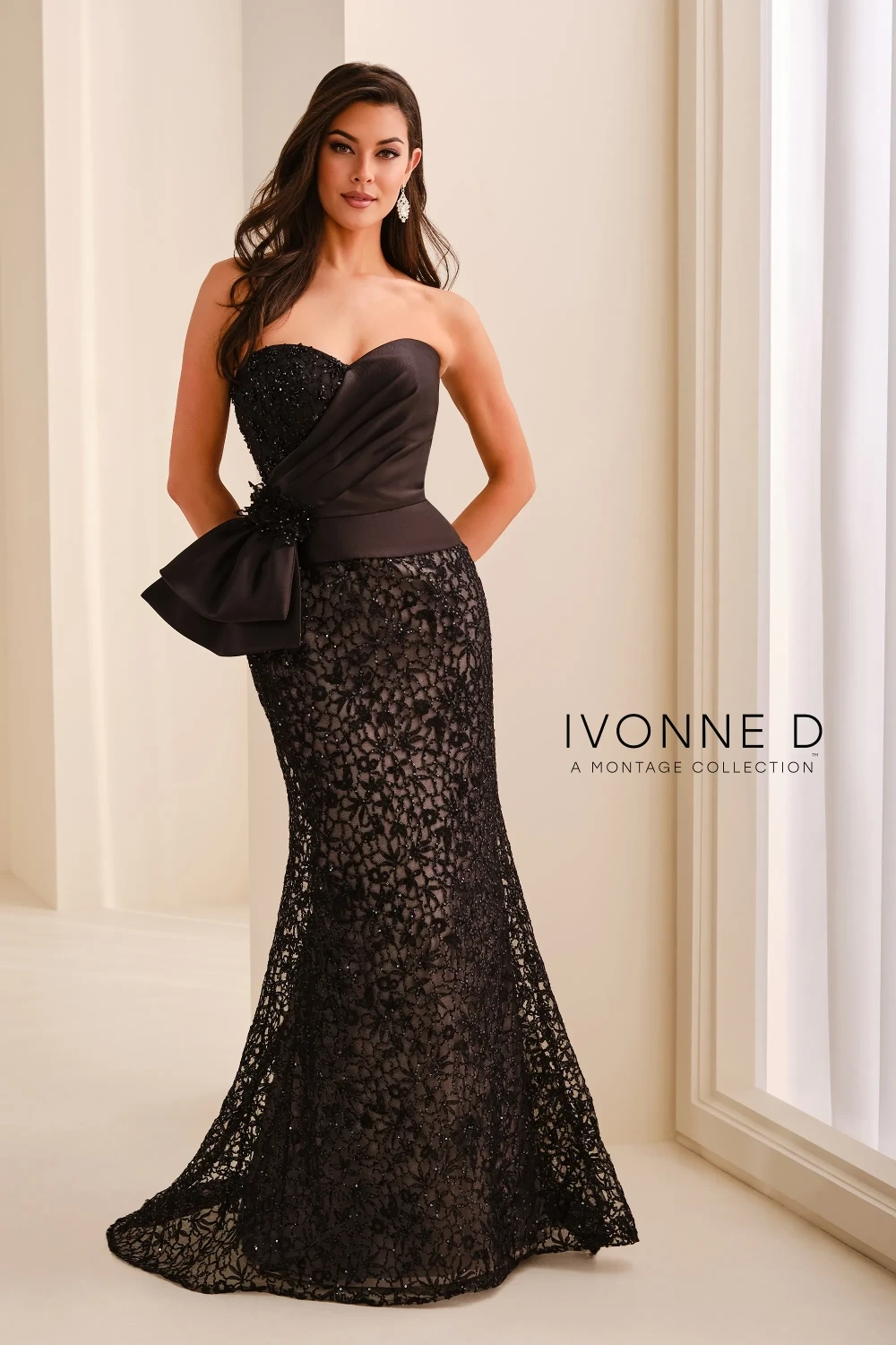 A model wearing a long, elegant black strapless gown with intricate lace and sequin detailing, a fitted silhouette, and a statement bow at the waist, standing against a neutral background.