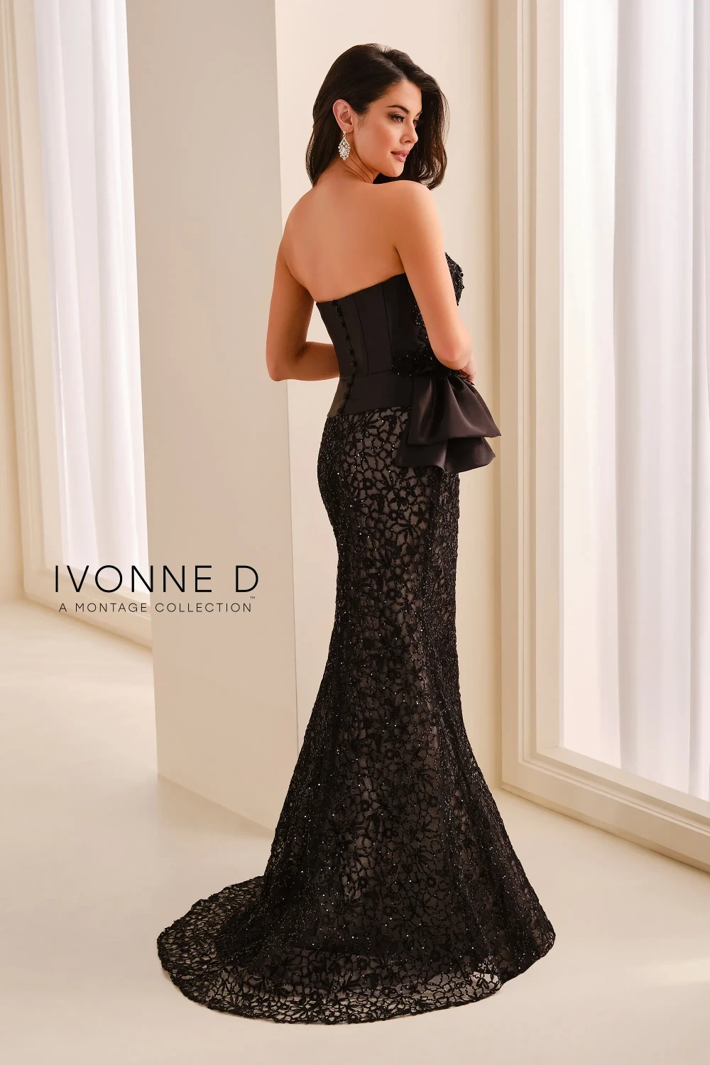 A model wearing a long, elegant black strapless gown with intricate lace and sequin detailing, a fitted silhouette, and a statement bow at the waist, standing against a neutral background.
