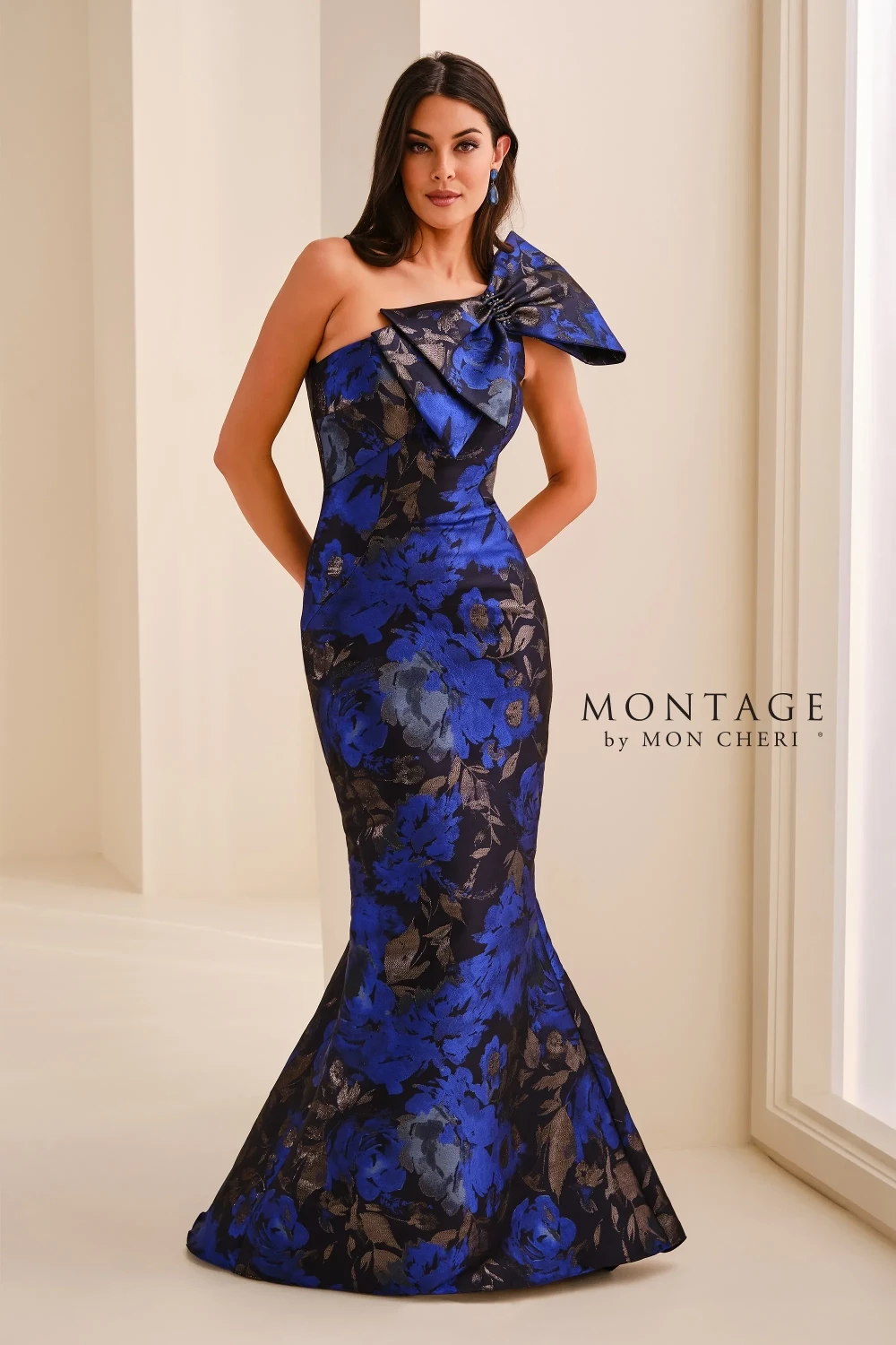 A model wearing a long, elegant royal blue and black floral one-shoulder gown with a dramatic bow detail, standing against a neutral background.