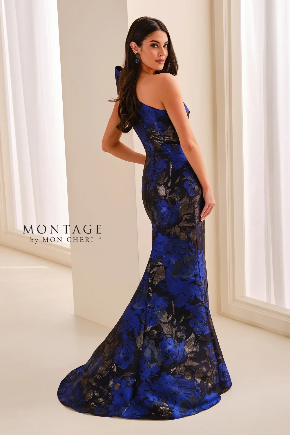 A model wearing a long, elegant royal blue and black floral one-shoulder gown with a dramatic bow detail, standing against a neutral background.