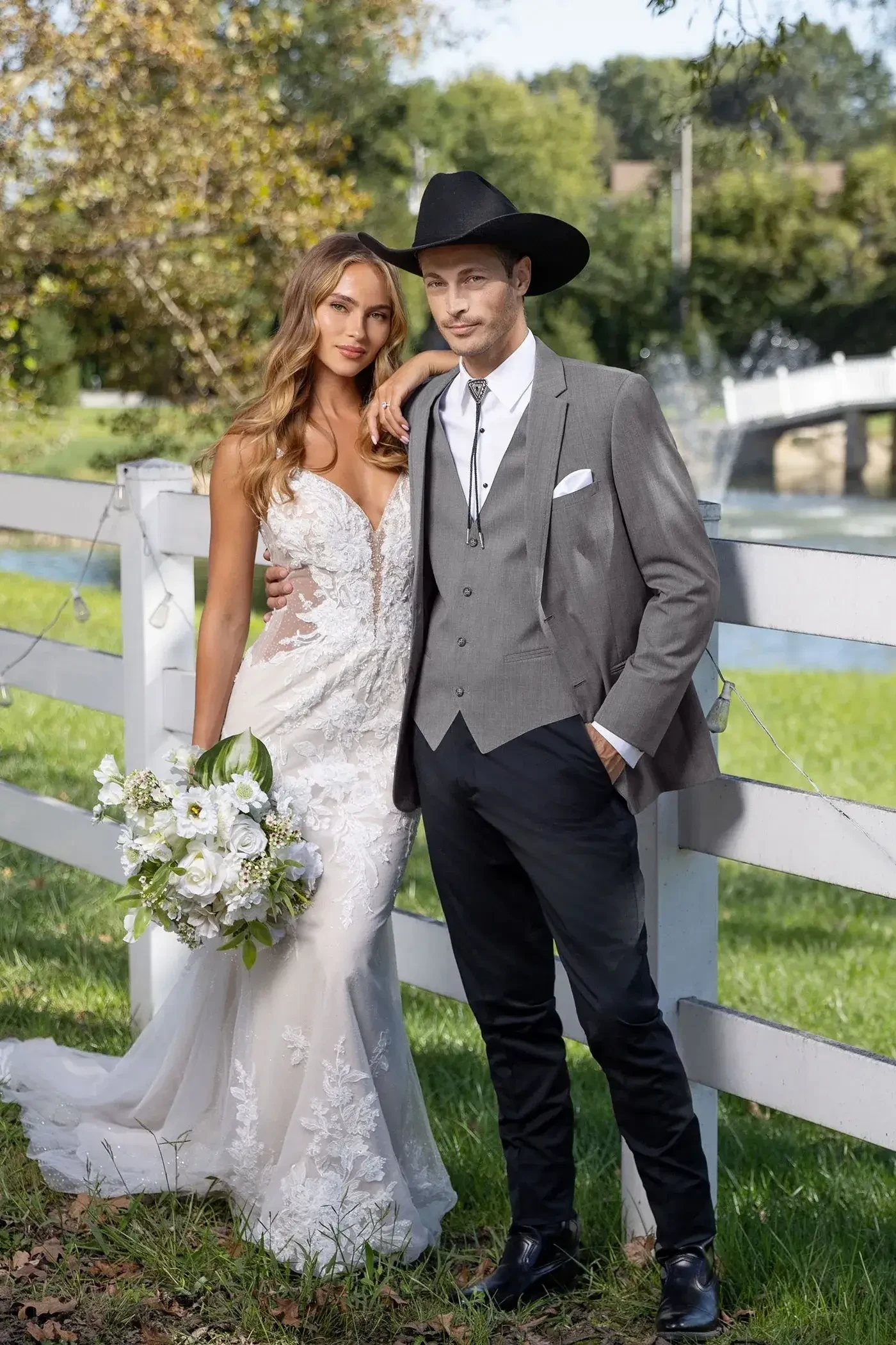 A couple poses together in a scenic outdoor setting, with the woman wearing an elegant wedding dress and holding a bouquet of flowers, while the man is dressed in a formal outfit complete with a cowboy hat. They stand beside a white fence with greenery in the background.