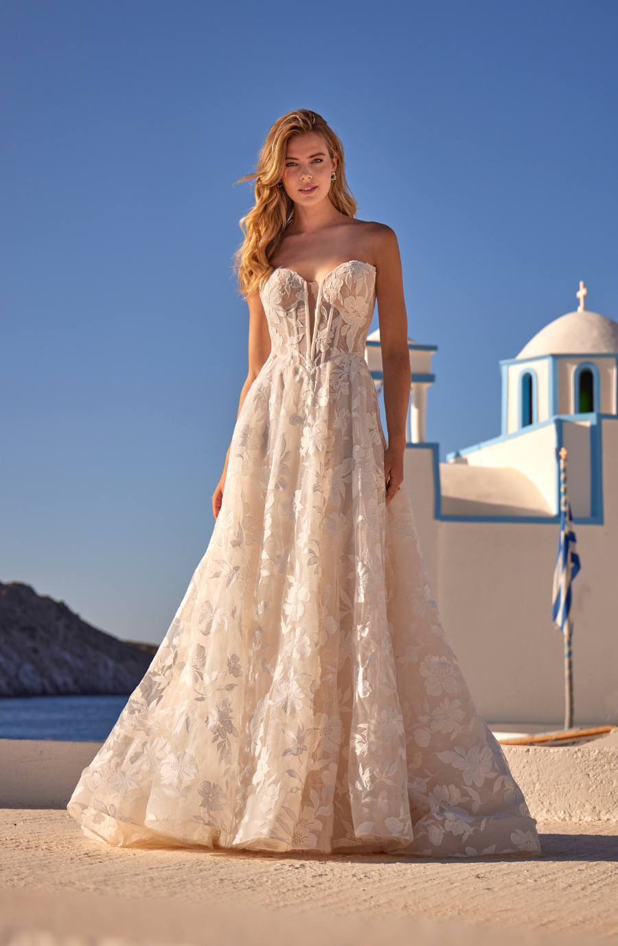 A woman in an elegant, strapless floral gown stands in a sunlit, coastal setting. A white church with a blue dome is in the background. The scene is serene and picturesque.